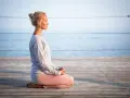 Kvinna med slutna ögon sitter i meditationsställning på en träbrygga vid havet. Hon utstrålar mental avslappning.