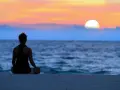 En person sitter i lotusställning på en strand och ser ut över havet vid solnedgången. En lugn stund för att meditera hemma, eller var som helst.