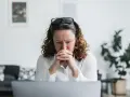 En kvinna med rufsigt hår och glasögon på huvudet sitter framför en laptop, djupt försjunken i tankar. Hennes ansiktsuttryck tyder på oro, kanske efter att ha utsatts för psykologisk misshandel.