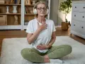 Kvinna med hörlurar sitter på en matta och utövar meditation och andning. Hennes ögon är slutna och hon har händerna på bröstet och magen.