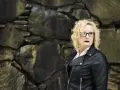 En kvinna med blont, lockigt hår och glasögon står framför en stenmur. Hon bär en svart läderjacka och mörkt läppstift. Hon utstrålar styrka, ett tecken på livet efter en destruktiv relation.