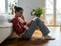 En ung tjej sitter på golvet, försjunken i sin telefon. Hennes hållning och ansiktsuttryck kan tyda på depression hos barn.