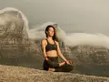 Kvinna med slutna ögon mediterar på en klippa med berg och moln i bakgrunden. Detta är en bild av vad betyder meditation.