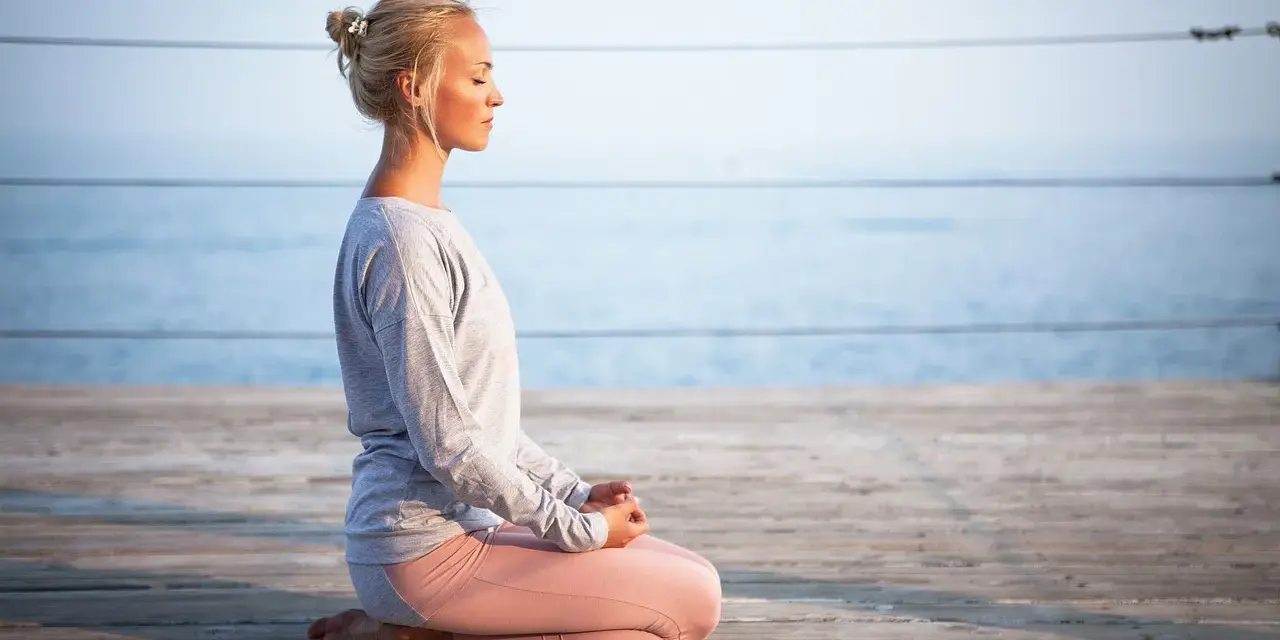Kvinna med slutna ögon sitter i meditationsställning på en träbrygga vid havet. Hon utstrålar mental avslappning.