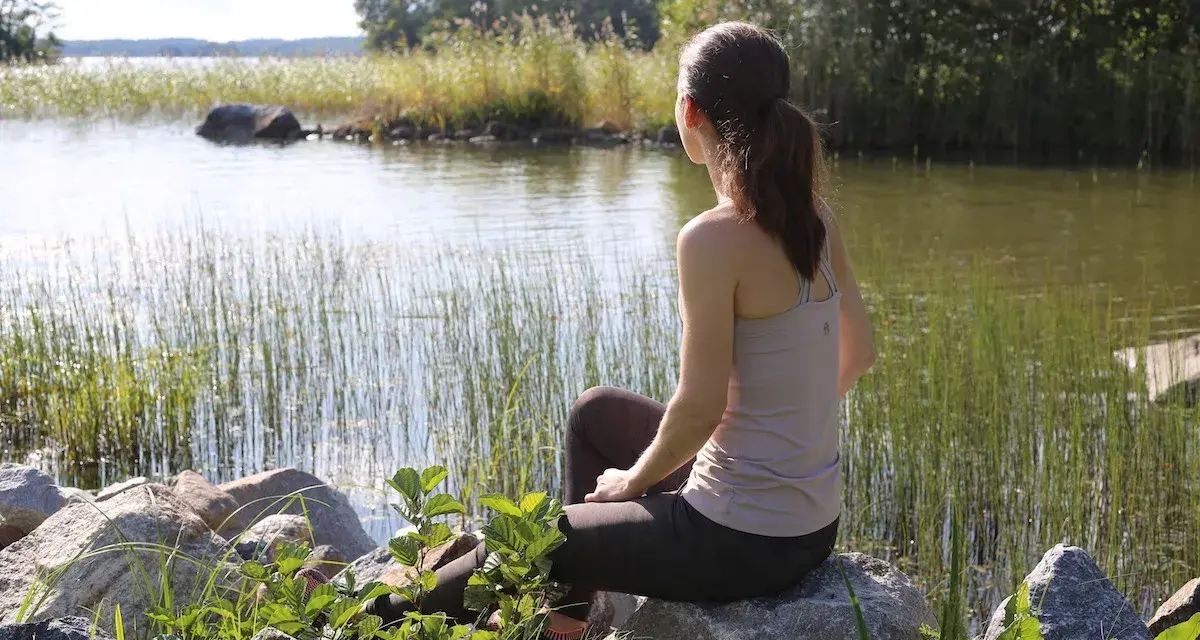 Kvinna sitter vid en sjö, omgiven av vass och stenar. En stund av avslappning mot ångest i naturens lugn.