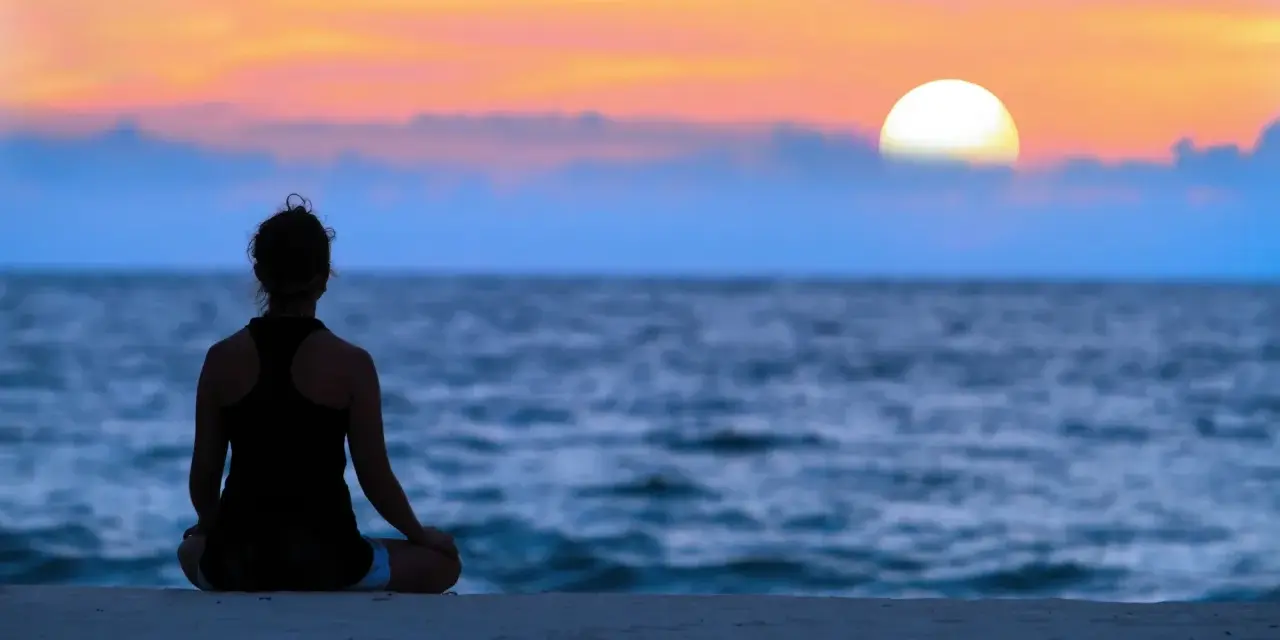 En person sitter i lotusställning på en strand och ser ut över havet vid solnedgången. En lugn stund för att meditera hemma, eller var som helst.