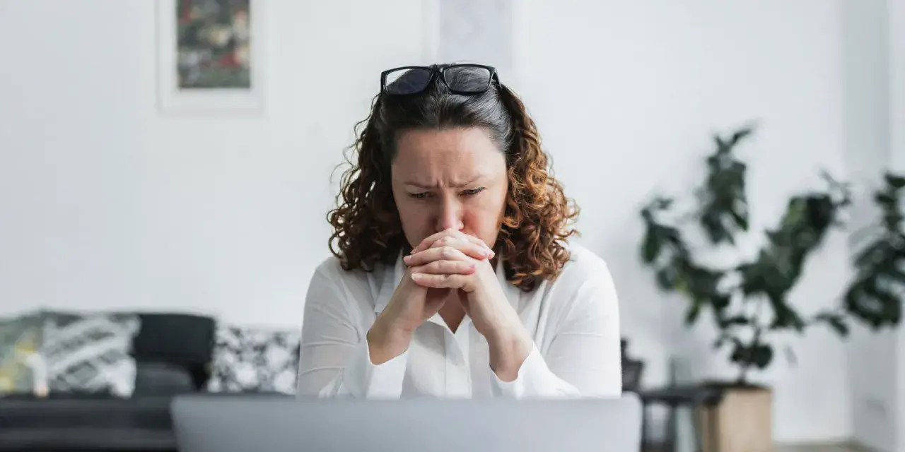 En kvinna med rufsigt hår och glasögon på huvudet sitter framför en laptop, djupt försjunken i tankar. Hennes ansiktsuttryck tyder på oro, kanske efter att ha utsatts för psykologisk misshandel.