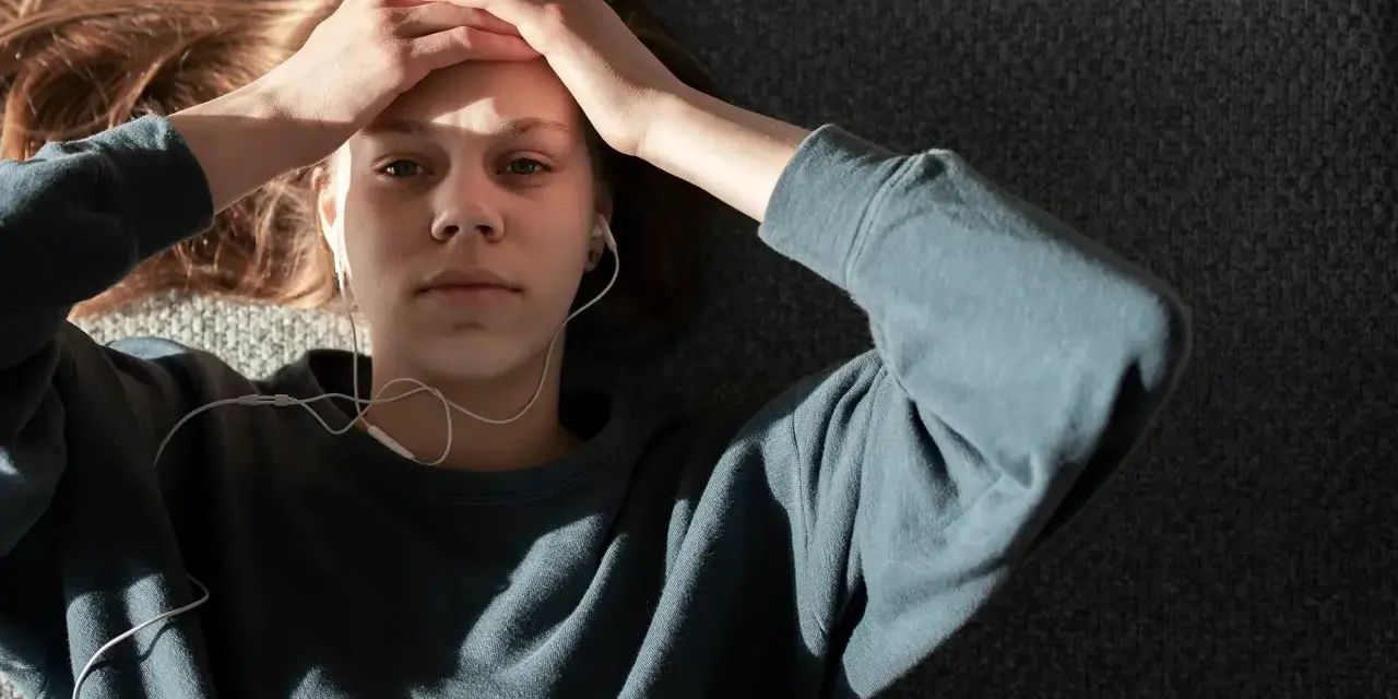 En ung person med hörlurar ligger på en grå yta, med händerna mot pannan. Kanske lyssnar hen på musik för att få bort ångest snabbt.