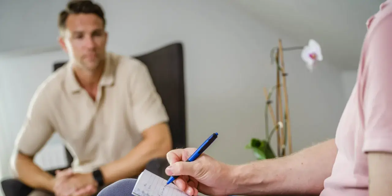 En person skriver anteckningar under ett samtal, kanske en del av act kbt-terapi. En annan person sitter i bakgrunden.