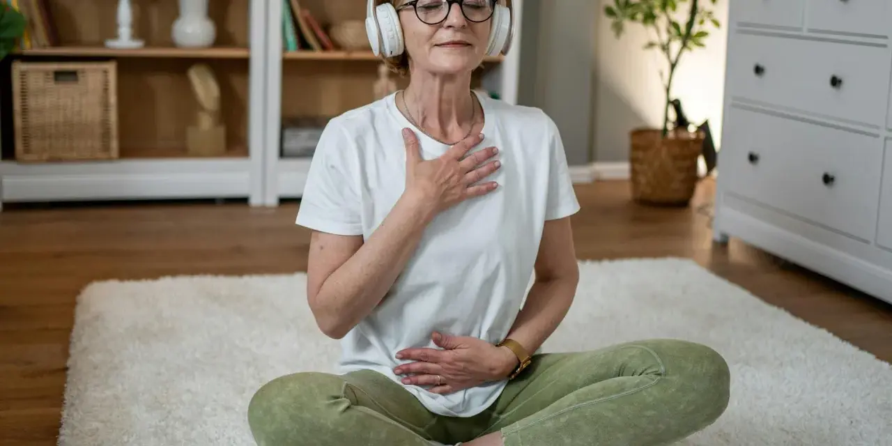 Kvinna med hörlurar sitter på en matta och utövar meditation och andning. Hennes ögon är slutna och hon har händerna på bröstet och magen.