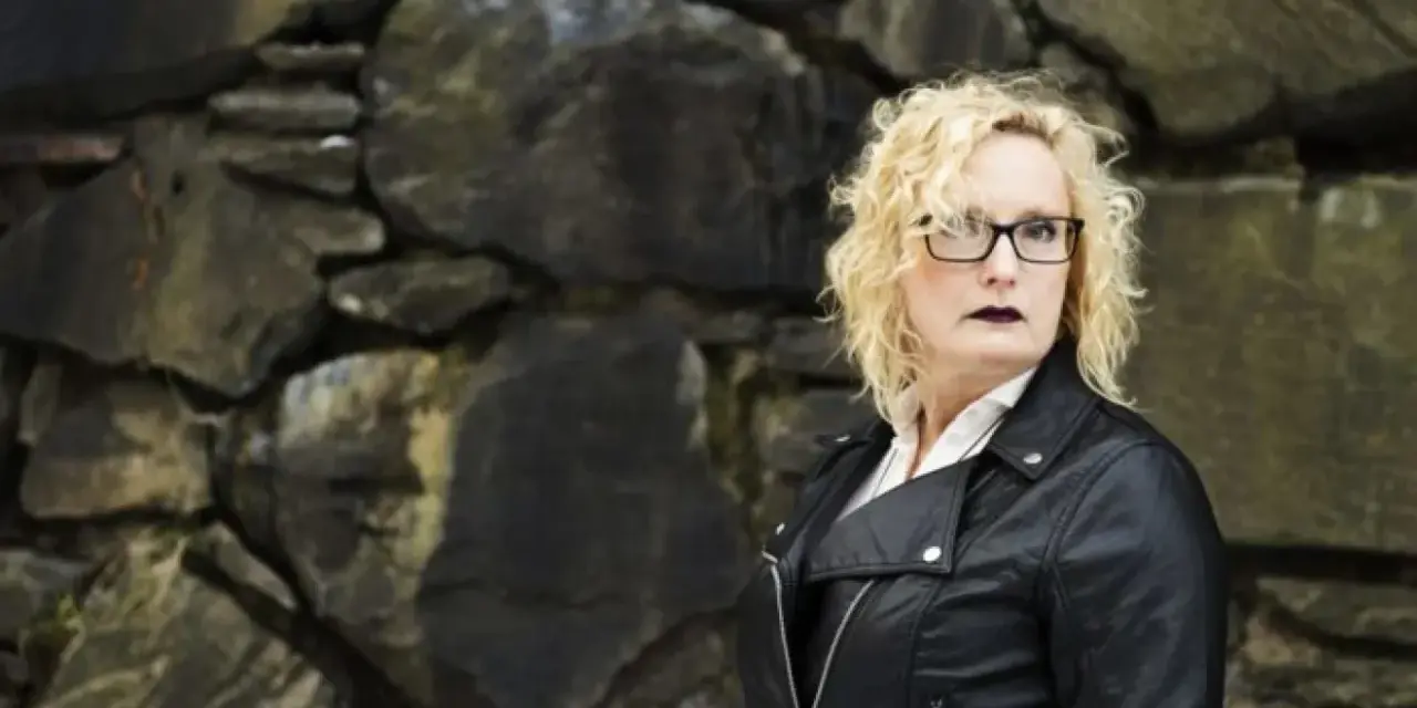 En kvinna med blont, lockigt hår och glasögon står framför en stenmur. Hon bär en svart läderjacka och mörkt läppstift. Hon utstrålar styrka, ett tecken på livet efter en destruktiv relation.