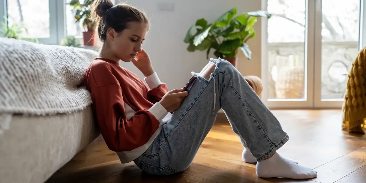 En ung tjej sitter på golvet, försjunken i sin telefon. Hennes hållning och ansiktsuttryck kan tyda på depression hos barn.