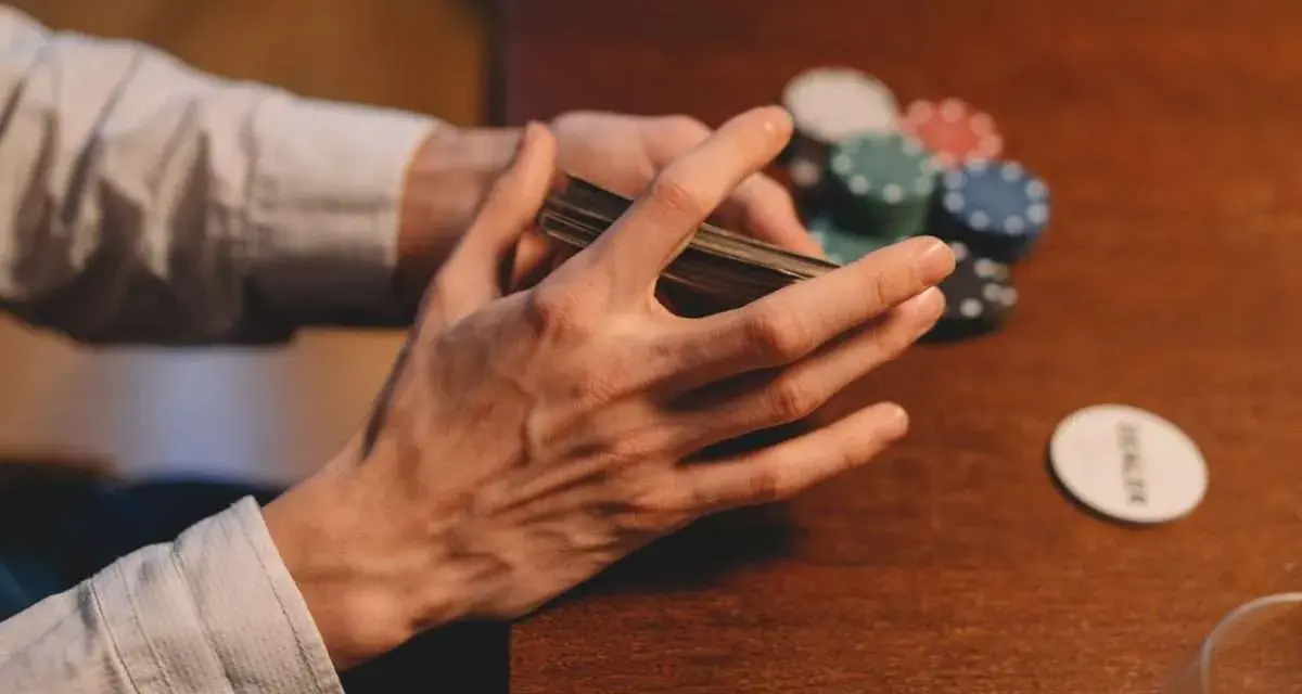 Händer som blandar kort vid ett pokerbord, med marker och en "dealer"-bricka. En påminnelse om att spelberoende behandling finns tillgänglig.