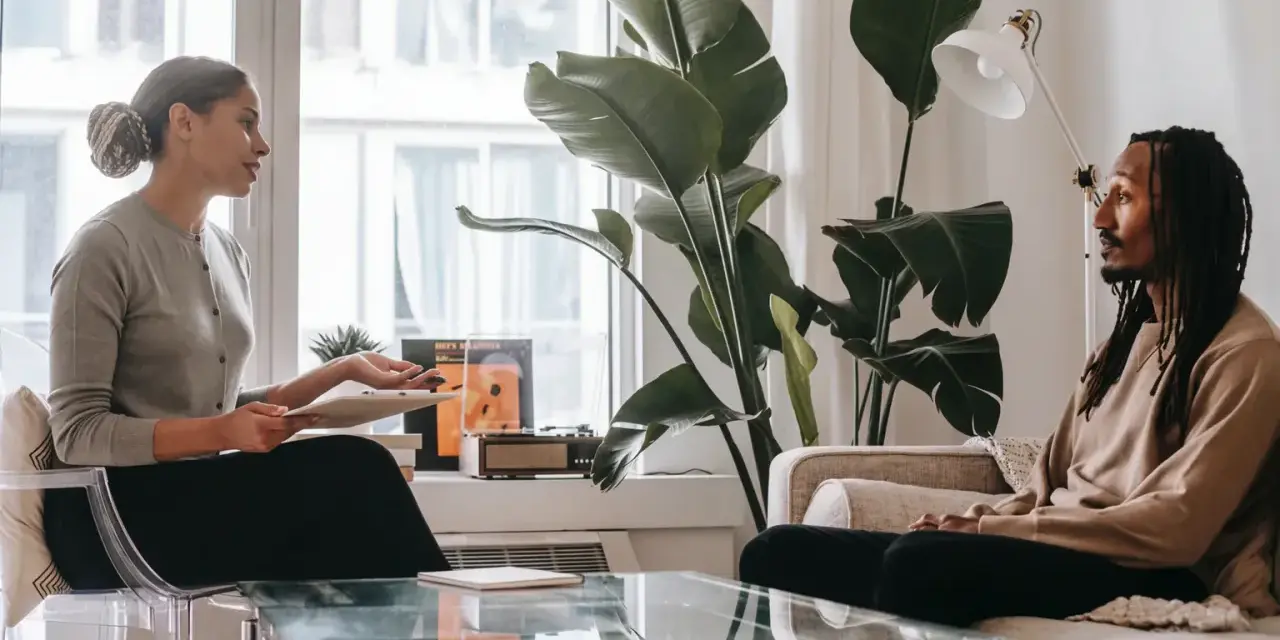 En kvinna och en man sitter mitt emot varandra i ett rum. Kvinnan håller en surfplatta och talar. Mannen lyssnar. Kanske diskuterar de hur mycket kostar terapi.