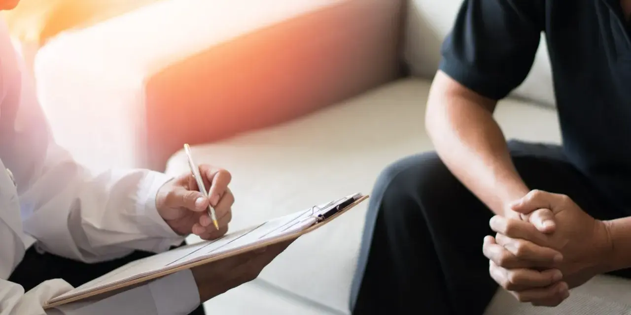 En terapeut skriver anteckningar medan en patient sitter med händerna knäppta. Bilden illustrerar vad betyder terapi – en plats för samtal och stöd.