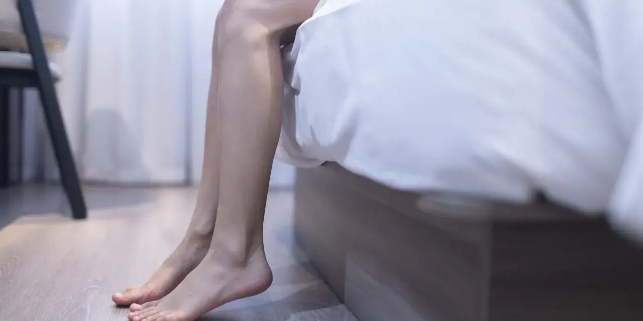 En person sitter på sängkanten, benen dinglar. Kanske upplever personen muskelryckningar vid vila, en vanlig men ibland oroande känsla.