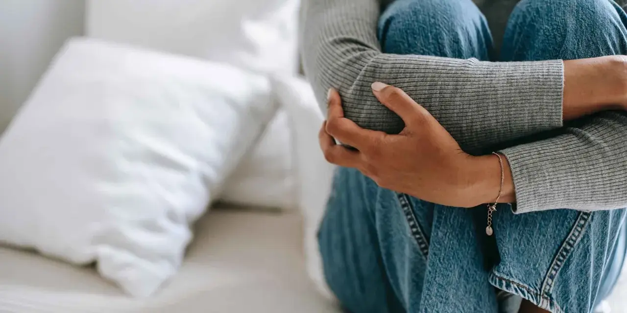 Person sitter hopkrupen på en säng, omfamnar sina knän. Klädd i jeans och grå tröja, utstrålar bilden extrem ångest.