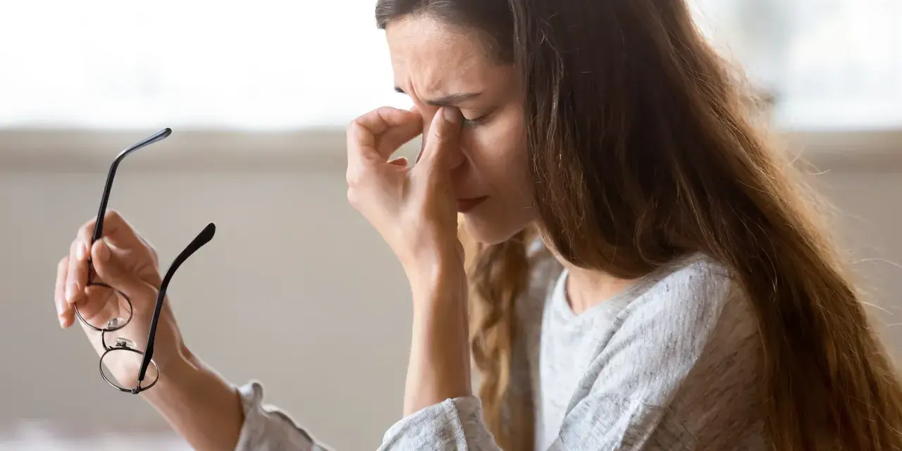 Kvinna med glasögon i handen, gnuggar sig i ögonen. Kanske tecken på muskelryckningar stress.