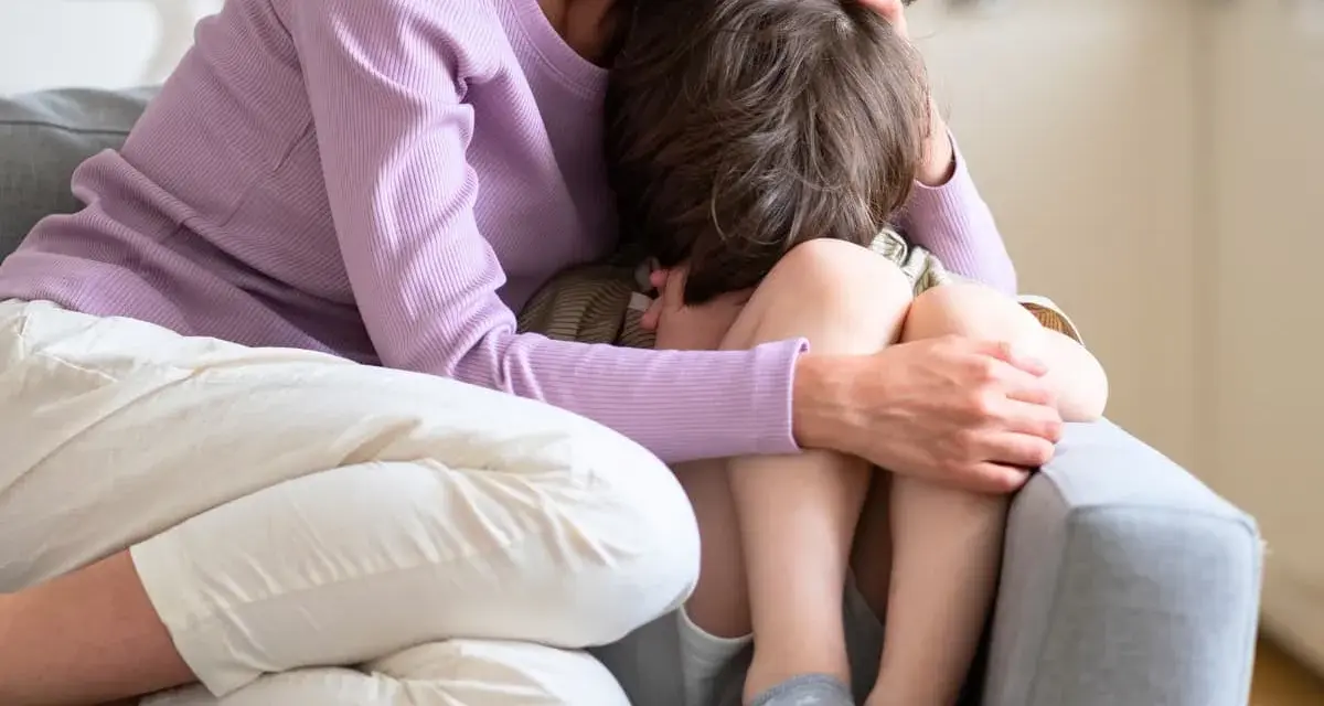En mamma tröstar sitt ledsna barn på en soffa. Hon håller om honom med kärlek.