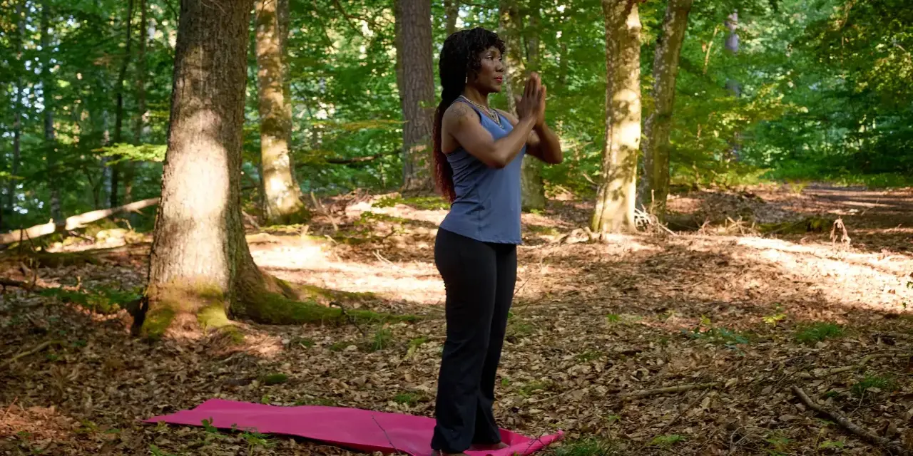 Kvinna i skogen på en yogamatta, med händerna i bön. En stund av avslappning och lugn för att minska stress.