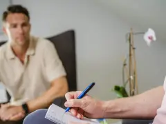 En person skriver anteckningar under ett samtal, kanske en del av act kbt-terapi. En annan person sitter i bakgrunden.
