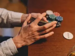 Händer som blandar kort vid ett pokerbord, med marker och en "dealer"-bricka. En påminnelse om att spelberoende behandling finns tillgänglig.