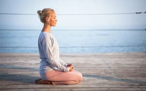 Kvinna med slutna ögon sitter i meditationsställning på en träbrygga vid havet. Hon utstrålar mental avslappning.