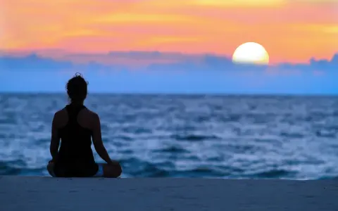 En person sitter i lotusställning på en strand och ser ut över havet vid solnedgången. En lugn stund för att meditera hemma, eller var som helst.