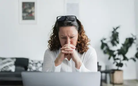 En kvinna med rufsigt hår och glasögon på huvudet sitter framför en laptop, djupt försjunken i tankar. Hennes ansiktsuttryck tyder på oro, kanske efter att ha utsatts för psykologisk misshandel.