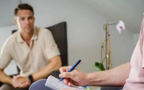 En person skriver anteckningar under ett samtal, kanske en del av act kbt-terapi. En annan person sitter i bakgrunden.