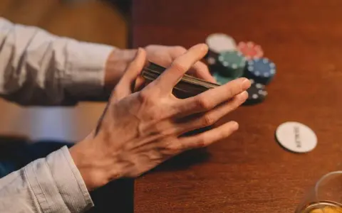 Händer som blandar kort vid ett pokerbord, med marker och en "dealer"-bricka. En påminnelse om att spelberoende behandling finns tillgänglig.