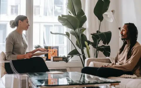 En kvinna och en man sitter mitt emot varandra i ett rum. Kvinnan håller en surfplatta och talar. Mannen lyssnar. Kanske diskuterar de hur mycket kostar terapi.