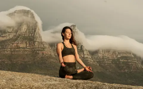 Kvinna med slutna ögon mediterar på en klippa med berg och moln i bakgrunden. Detta är en bild av vad betyder meditation.