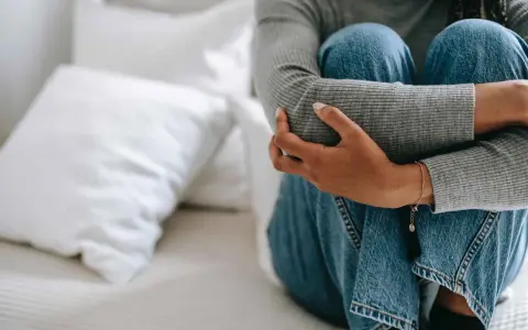 Person sitter hopkrupen på en säng, omfamnar sina knän. Klädd i jeans och grå tröja, utstrålar bilden extrem ångest.