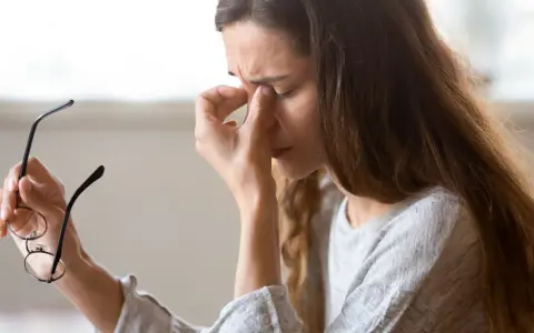 Kvinna med glasögon i handen, gnuggar sig i ögonen. Kanske tecken på muskelryckningar stress.