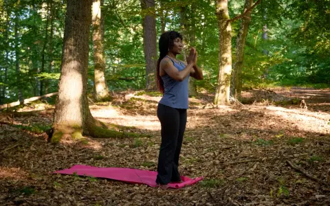 Kvinna i skogen på en yogamatta, med händerna i bön. En stund av avslappning och lugn för att minska stress.