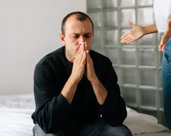 En man sitter med slutna ögon och händerna knäppta, uppenbart ledsen. En kvinna står bakom honom och gestikulerar, vilket antyder en destruktiv relation.