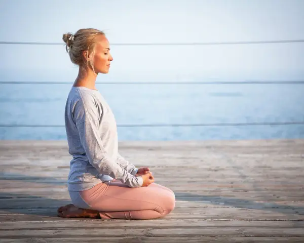 Kvinna med slutna ögon sitter i meditationsställning på en träbrygga vid havet. Hon utstrålar mental avslappning.