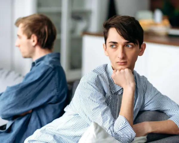 Två män sitter på en soffa, vända bort från varandra. Den ena har armarna i kors, den andra vilar hakan i handen. Deras kroppsspråk antyder ett grandiost beteende, kanske en tyst konflikt.