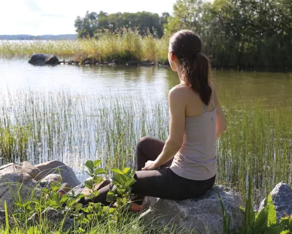 Kvinna sitter vid en sjö, omgiven av vass och stenar. En stund av avslappning mot ångest i naturens lugn.