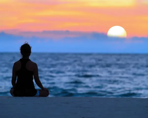 En person sitter i lotusställning på en strand och ser ut över havet vid solnedgången. En lugn stund för att meditera hemma, eller var som helst.