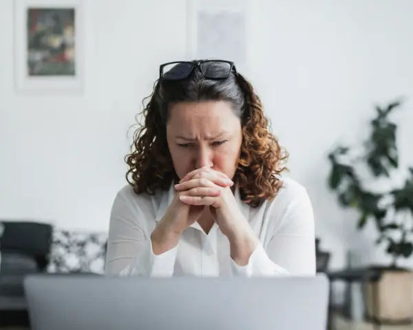 En kvinna med rufsigt hår och glasögon på huvudet sitter framför en laptop, djupt försjunken i tankar. Hennes ansiktsuttryck tyder på oro, kanske efter att ha utsatts för psykologisk misshandel.