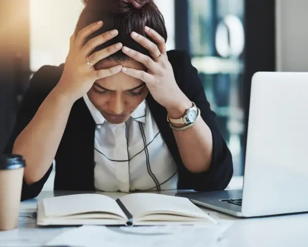 En kvinna med händerna i håret sitter vid ett skrivbord med en öppen anteckningsbok och en laptop. Hon ser stressad ut, vilket påverkar trivseln på jobbet.