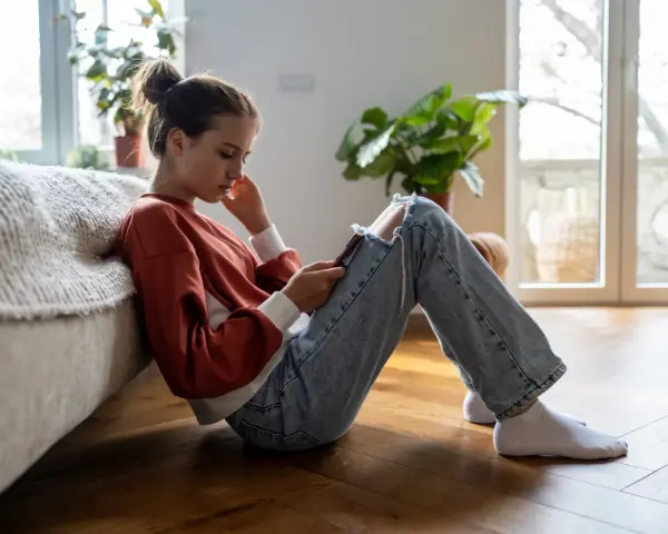 En ung tjej sitter på golvet, försjunken i sin telefon. Hennes hållning och ansiktsuttryck kan tyda på depression hos barn.