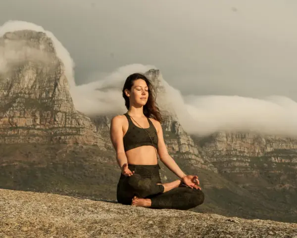 Kvinna med slutna ögon mediterar på en klippa med berg och moln i bakgrunden. Detta är en bild av vad betyder meditation.