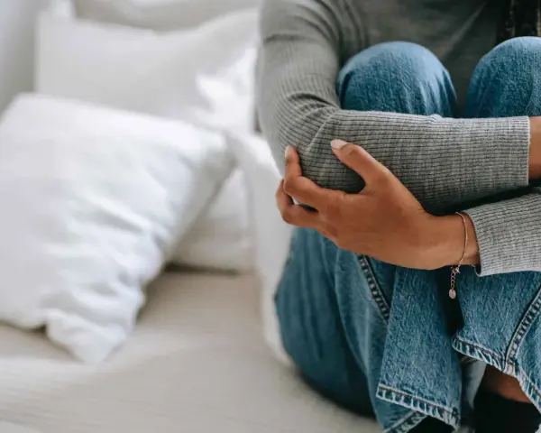 Person sitter hopkrupen på en säng, omfamnar sina knän. Klädd i jeans och grå tröja, utstrålar bilden extrem ångest.