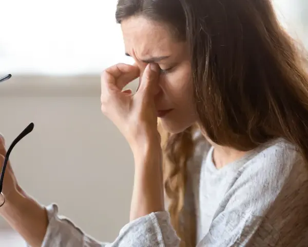 Kvinna med glasögon i handen, gnuggar sig i ögonen. Kanske tecken på muskelryckningar stress.