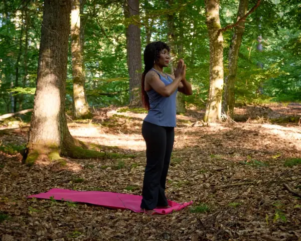 Kvinna i skogen på en yogamatta, med händerna i bön. En stund av avslappning och lugn för att minska stress.