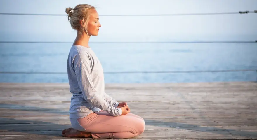 Kvinna med slutna ögon sitter i meditationsställning på en träbrygga vid havet. Hon utstrålar mental avslappning.