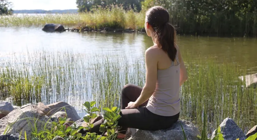 Kvinna sitter vid en sjö, omgiven av vass och stenar. En stund av avslappning mot ångest i naturens lugn.